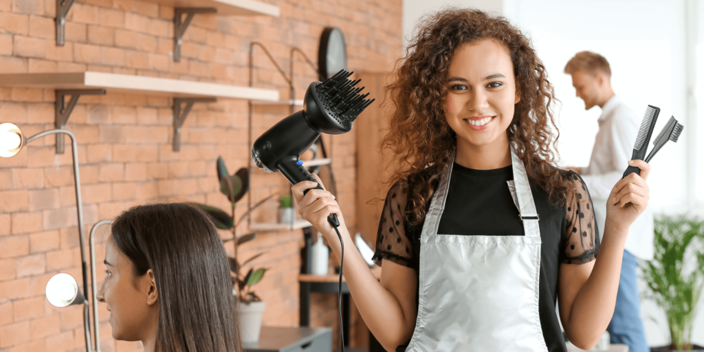 Smiling hairdresser holding a hairdryer and combs in a modern salon, ready to begin a client’s styling session.
