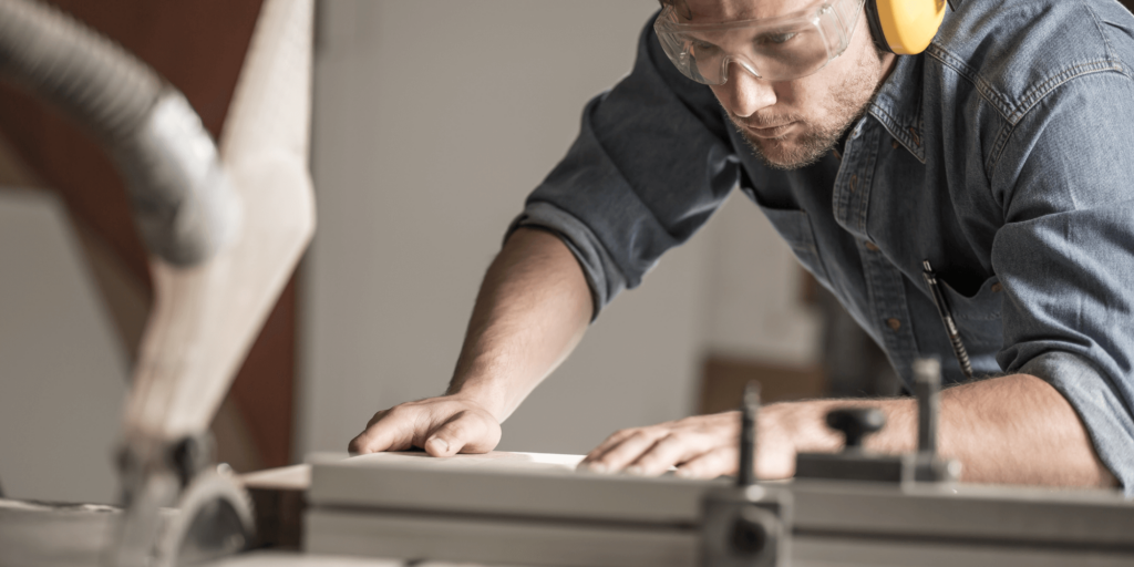 Joiner measuring timber in a workshop with power tools and wooden panels in the background.