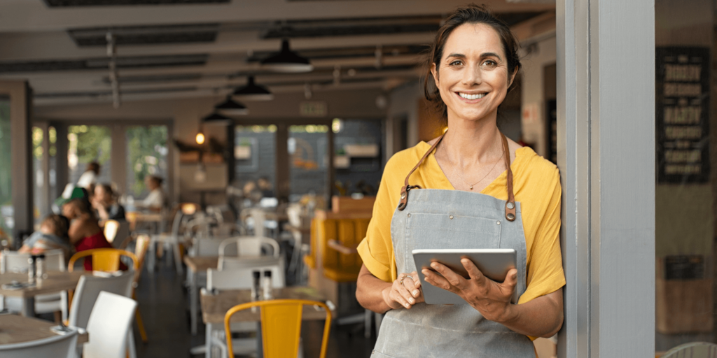 Smiling restaurant owner wearing a yellow top and apron, holding a digital tablet while standing at the entrance of a modern café.
